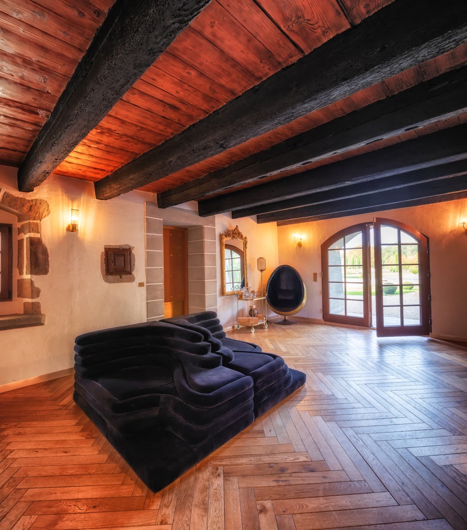 Grand salon à poutres anciennes avec canapé modulaire de velours et portes-fenêtres cintrées